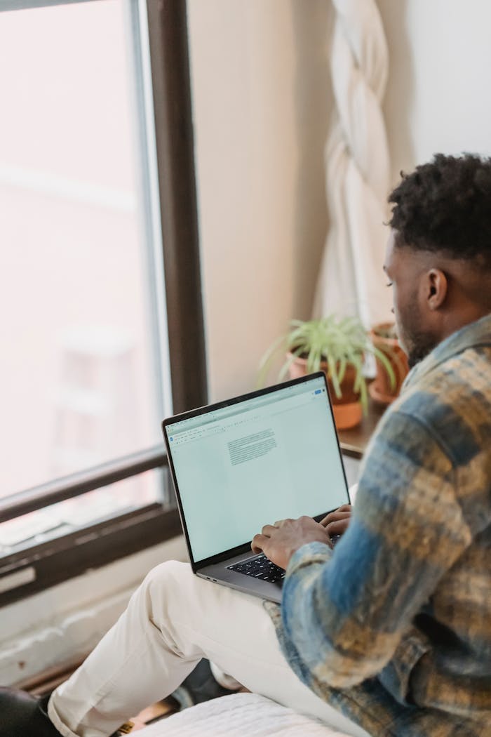 About Back view of African American male in casual clothes sitting in cozy room in daylight and typing information on computer keyboard