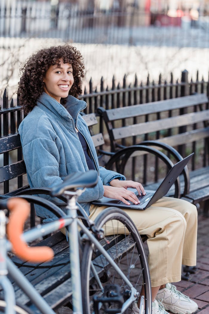 Services Positive young ethnic female freelancer with curly hair in trendy warm jacket smiling and looking at camera while browsing laptop sitting on wooden bench in city park
