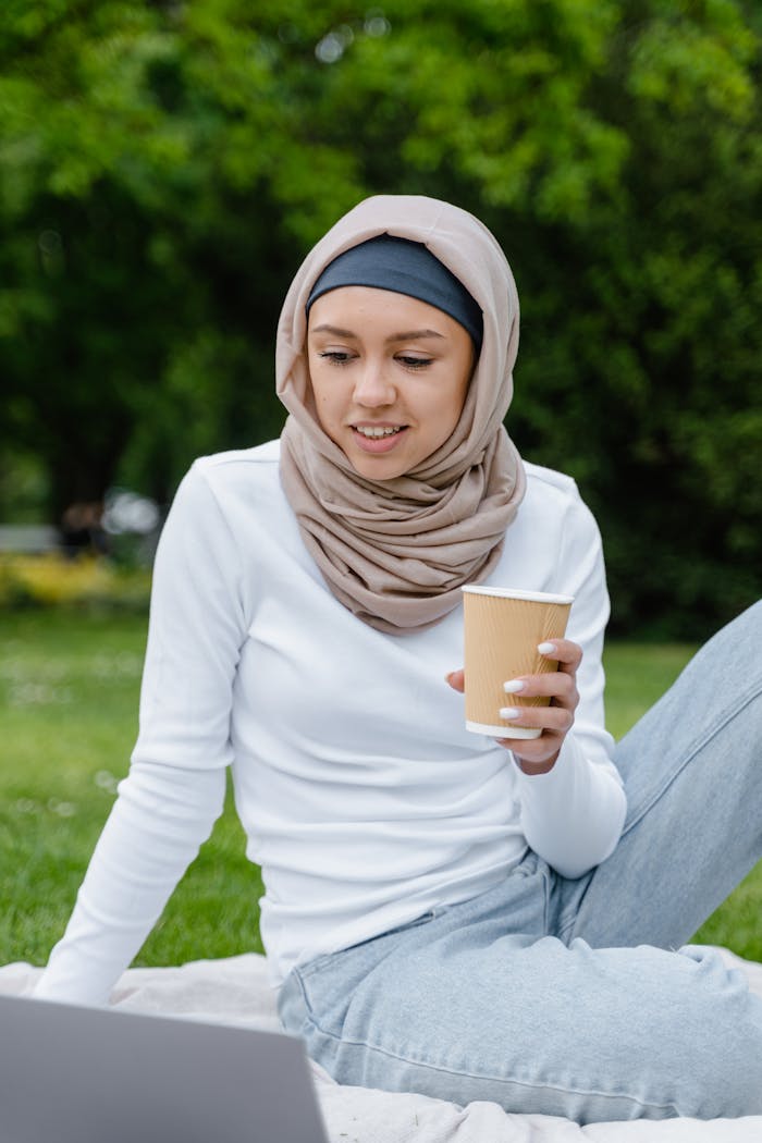 A Muslim woman in a hijab enjoying a coffee in a park while using a laptop. Outdoor setting with greenery.